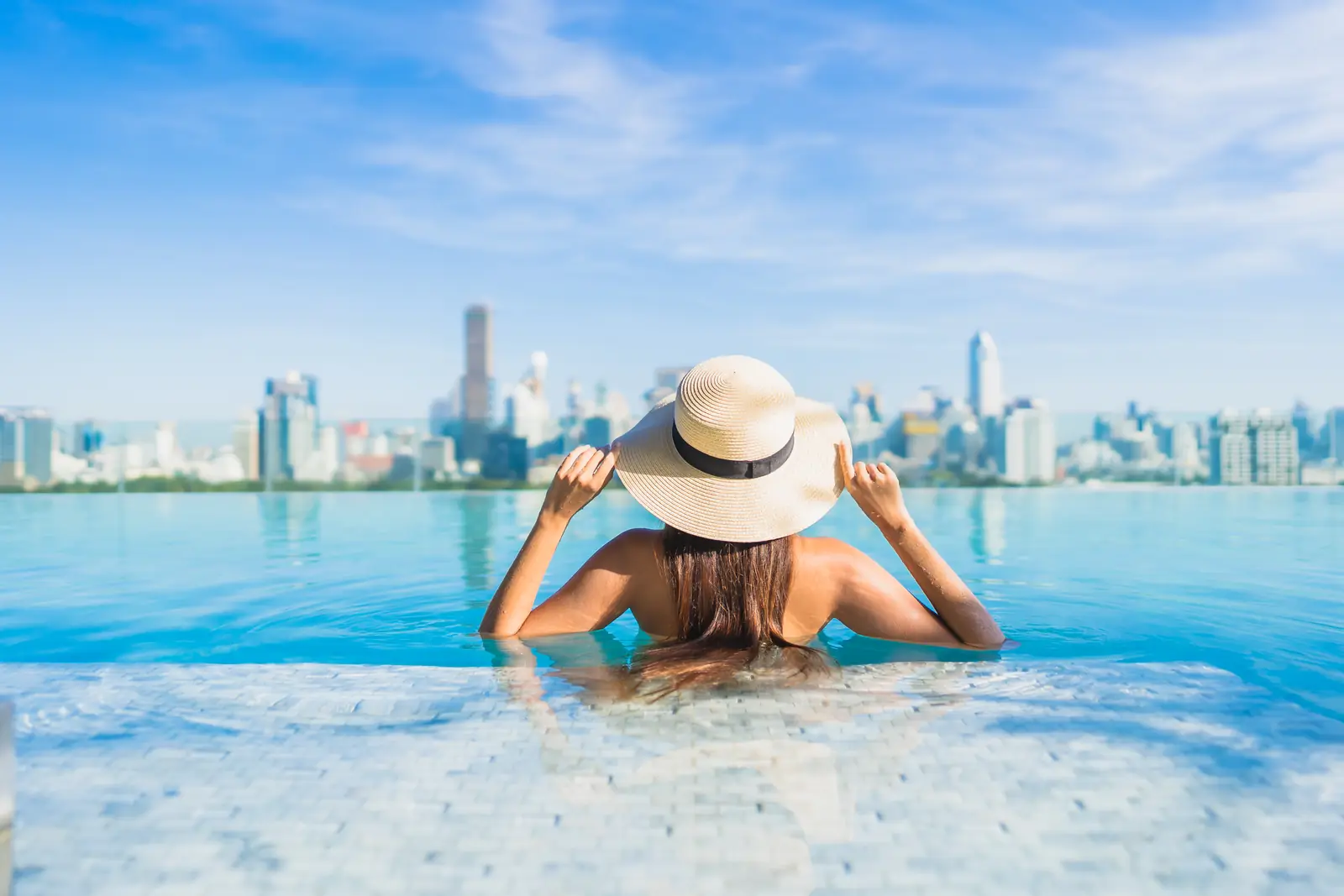A woman in a sun hat relaxing in a rooftop infinity pool above the city